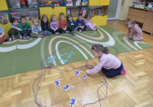 Dzieci siedzą na dywanie, dziewczynka liczy obrazki dinozaurów.