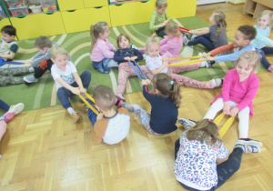 Dzieci siedzą na podłodze w parach na przeciwko siebie i trzymają szarfy.