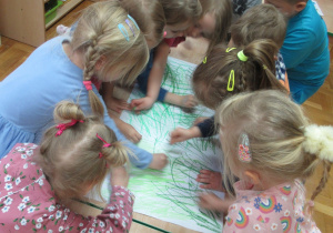 Dzieci stoją przy stole i rysują łąkę.