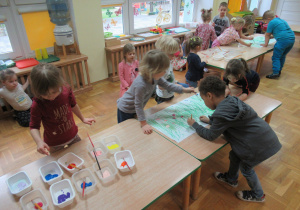 Dzieci stoją przy stołach i malują farbami.