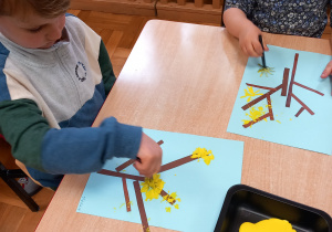 Dzieci siedzą przy stolikach i malują farbami.