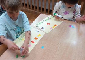 Dzieci wyklejają kartkę kolorowymi kwiatkami.