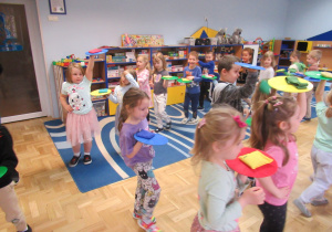 Dzieci maszeruja po sali, w ręku trzmaja krążek gimnastyczny, na którym leży woreczek lub szarfa.