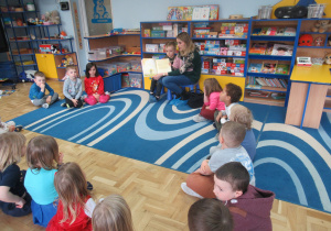 Mama chłopca czyta dzieciom książkę, dzieci siedzą w kole na podłodze.