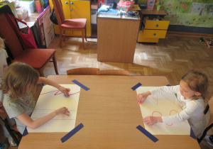Dziewczynki siedzą przy stole i przesuwają nakrętki po liniach narysowanych na kartce papieru.