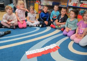 Dzieci siedzą na dywanie, przed nimi kartki białe i czerwone z literą f.