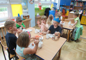 Dzieci siedzą przy stolikach, jedzą urodzinowy poczęstunek.