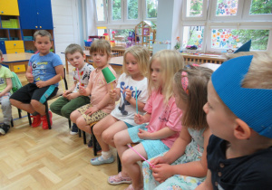Dzieci siedzą na krzesełkach i podają sobie kubek za pomocą słomek.