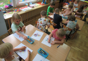 Dzieci siedzą przy stolikach i kolorują.