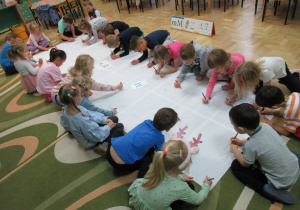 Dzieci siedzą wokół dużego białego kartonu, na którym piszą litery M.