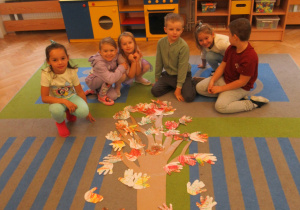 Dzieci pozują do zdjęcia wokół drzewa ułożonego na dywanie.