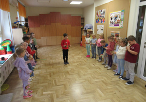 Dzieci stoją w dwóch szeregach, między nimi chłopiec. Wszystkie dzieci grają na instrumentach.