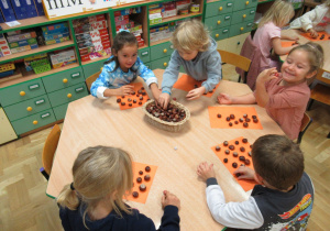 Dzieci siedzą przy stolikach i układają kasztany na kartkach.