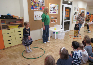 Dzieci siedzą na dywanie, przed nimi panie i 3 dzieci stoi w obręczach podczas zajęć o orientacji przestrzennej.