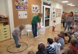 Pani i chłopiec stoją i wskazują rękami podłogę, pozostałe dzieci siedzą na podłodze.