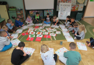 Dzieci siedzą w kole podczas zajęć, przed dziećmi leżą książki, na środku ilustracje przedstawiające warzywa.