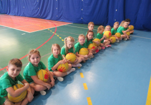 Dzieci siedzą na podłodze, trzymają w rękach zółte piłki podszas zajęć sportowych w szkole.