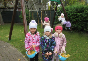 Dwie dziewczynki huśtają się na huśtawce. Trzy dziewczynki pozują do zdjęcia, trzymają w rękach wiaderka, w których są jesienne liście.