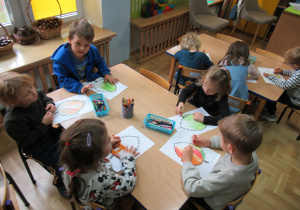 Dzieci siedzą przy stolikach i kolorują kredkami szablony liści.