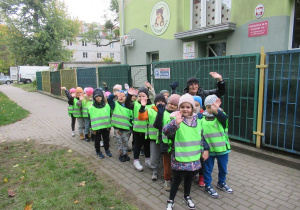 Dzieci ubrane w odblaskowe kamizelki stoją ustawione w pary przed przedszkolem.