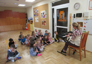 Dzieci siedzą na podłodze przed panią grającą na pianinie.