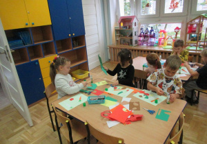 Dzieci siedzą przy stolikach, wycinają i przyklejają warzywa z papieru.