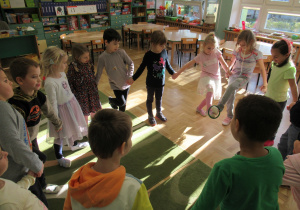 Dzieci stoją w kole trzymając się za ręce, dziewczynka przekazuje obręcz drugiej dziewczynce stopą.e