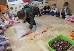 Dzieci siedzą w kole, chłopiec układa na podłodze rytm z kasztanów i liści.
