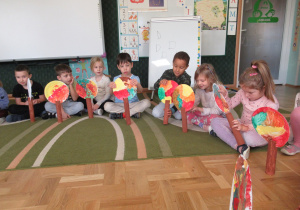 Dzieci siedzą na dywanie, trzymają prace plastyczne: drzewa z rolek i talerzyków.