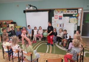 Dzieci siedzą na krzesełkach ustawionych w koło, chłopiec stoi w środku i trzyma w ręku balonik.