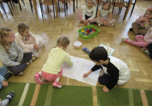 Dzieci siedzą na podłodze. Dziewczynka i chłopiec wodzą polcem po cyfrze 6 napisanej na kartce.