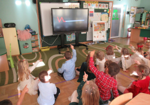 Dzieci siedzą na dywanie, przed nimi tablica multimedialna, dzieci śledzą kształty pojawiające się na ekranie za pomocą rąk w rytm muzyki.