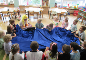 Dzieci siedzą w kole i trzymają niebieski materiał.