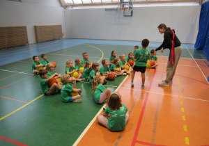 Dzieci siedzą na podłodze, każdy trzyma piłkę do koszykówki.