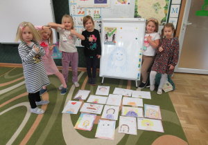 Dziewczynki stoją wokół ułożonych na dywanie prac plastycznych - Portret z uśmiechem.
