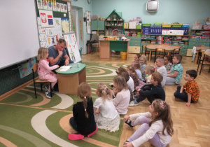 Dziewczynka z tatą siedzą przy stoliku, tata czyta książkę, pozostałe dzieci siedzą na dywanie i słuchają.