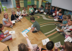 Dzieci siedzą w kole i trzymją w rękach kartki z literami.