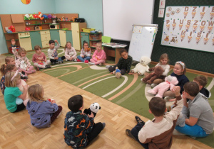 Dzieci siedzą w kole na dywanie i trzymają na kolanach swoje pluowe misie.