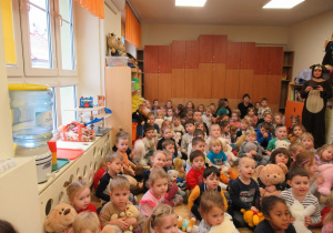 Dzieci z całęgo przedszkola siedzą na podłodze i trzymają swoje pluszowe misie, wśród nich pani przebrana za misia.