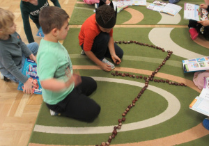 Dzieci układają na dywanie cyfrę 7 z kasztanów.