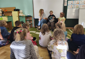 Dziewczynka z mamą i bratem siedzą na krzesełkach przed dziećmi. Chłopiec czyta dzieciom książkę o Elfach.