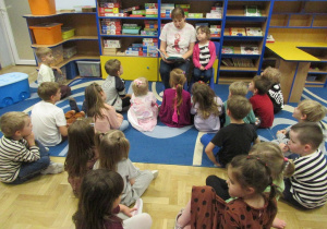 Dziewczynka z mamą siedzą na krzesełkach, mama czyta książkę, pozostałę dzieci siedzą na dywanie i słuchają.