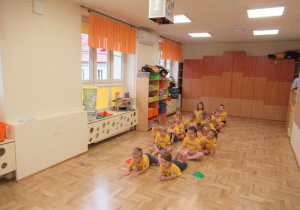 Dzieci siedzą w dwóch rzędach na podłodze podczas zajęć gimnastycznych.