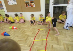 Dzieci siedzą w szeregu, chłopiec pokonuje tor przeszkód.