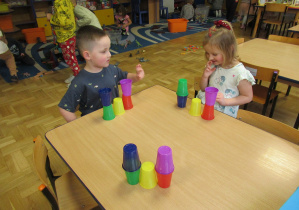 Dziewczynka i chłopiec siedzą przy stoliku i układają kolorowe kubeczki wg wzoru.