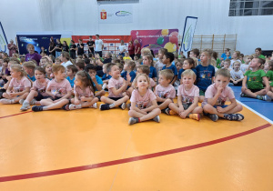 Dzieci siedzą na podłodze w sai gimnastycznej.