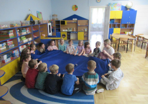 Dzieci siedzą w kole na dywanie, trzymają niebieski materiał podczas zajęć Roar Dance.