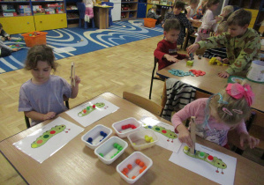 Dziewczynki siedzą przy stoliku i układają pomponiki wg wzory przy pomocy drewnianych szczypiec.