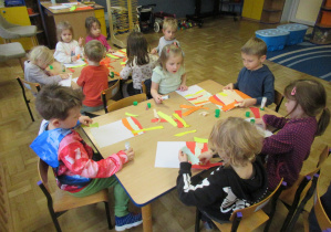 Dzieci siedzą przy stolikach i wyklejają kartki kolorowym papierem.