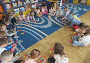 Dzieci siedzą w kole na dywanie, przed nimi figury geometryczne.